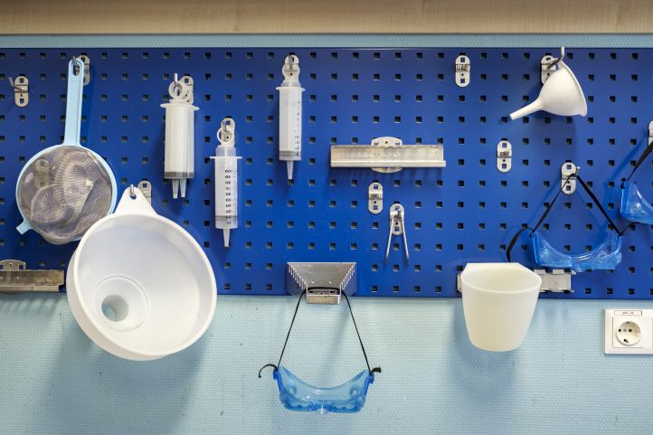 Laborausrüstung hängt an einer blauen Wand, darunter Spritzen, ein Sieb, Trichter, Schutzgläser, Schutzbrillen und Becher