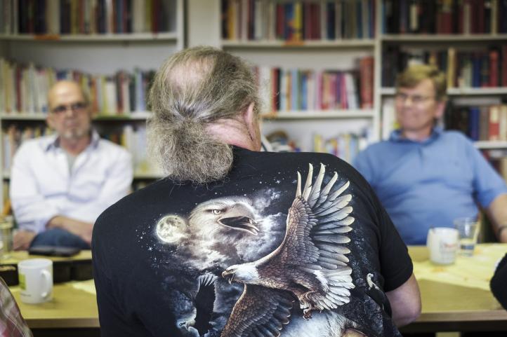 Ein Mann mit grauen Haaren und Zopf sitzt mit dem Rücken zum Betrachter an einem Tisch vor einem Bücherregal und andere hören ihm zu