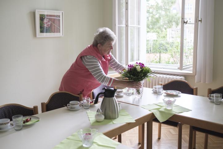 EIne ältere Frau bereitet einen Kaffeetisch für Gäste vor und steckt gerade Blumen in eine Vase