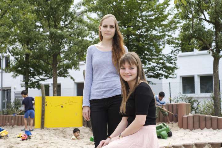 Zwei junge Frauen sitzen auf einem Spielplatz mit Kindern im Hintergrund und schauen lächelnd in die Kamera