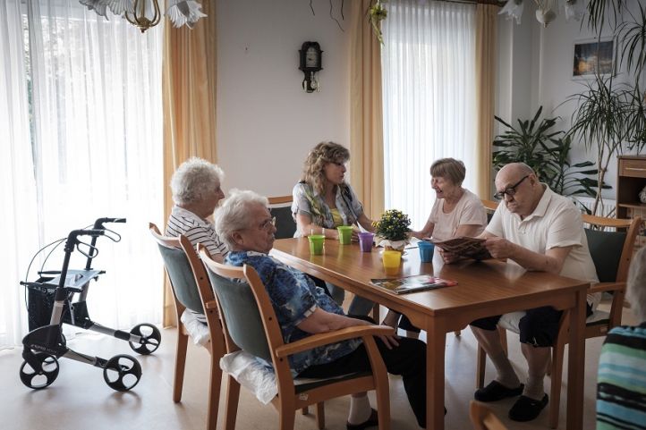 Ältere Bewohner*innen einer Pflegeeinrichtung sitzen um einen Tisch im Hintergrund ein Rollator