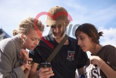Zwei junge Frauen und ein junger Mann stehen draußen und schauen auf ein Handy Smartphone